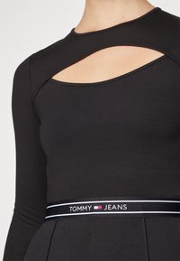 Maglietta nera a maniche lunghe con un'apertura sul collo ed un design aderente. Presenta un elastico nero con il logo "TOMMY JEANS" in bianco e rosso.