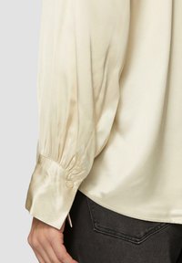 Blouse en satin crème avec des manches volumineuses, poignets boutonnés et une texture lisse, présentant de subtils plis le long des manches.
