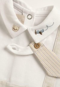 Polo en tissu blanc avec une broderie de ours beige, des détails brodés bleu et marron, et une sangle rayée beige clair avec un bouton en bois.