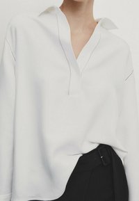 Camicia bianca a maniche lunghe con colletto a placca, caratterizzata da dettagli di cucitura nera lungo le cuciture e una vestibilità ampia, abbinata a pantaloni scuri.