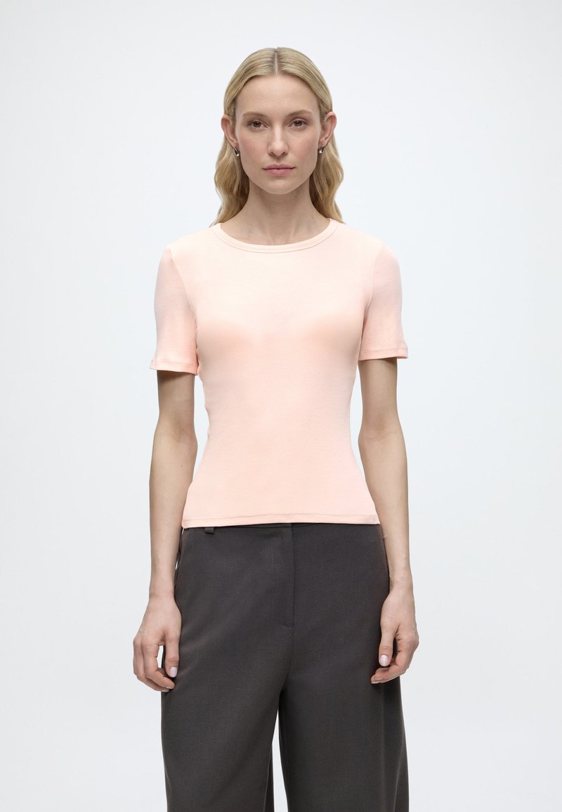 Ξανθιά γυναίκα που φοράει μια ανοιχτορόζ κοντομάνικη εφαρμοστή μπλούζα και φαρδιά σκούρα γκρι παντελόνια, στέκεται μπροστά σε ένα απλό λευκό φόντο.