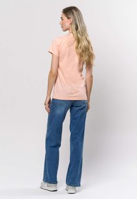 Hellpfirsichfarbenes kurzärmeliges T-Shirt mit Rundhalsausschnitt, kombiniert mit lockeren blauen Jeans. Sichtbare Nahtdetails, glatte Stoffstruktur und lässige Passform.