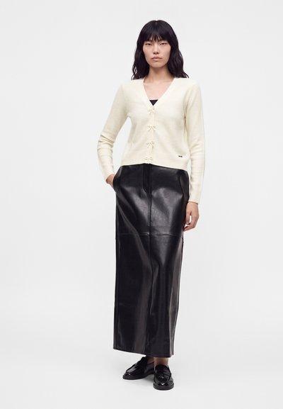 Femme portant un cardigan crème boutonné, une longue jupe en cuir noire et des mocassins noirs, debout avec une main dans la poche.