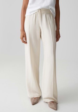 Personne portant un pantalon crème taille haute à jambes larges avec une ceinture élastique et un cordon de serrage, associé à des chaussures beige à bout pointu et un haut blanc.