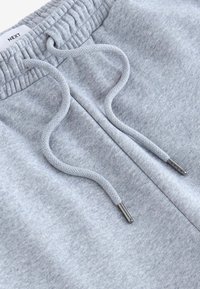 Graue Jogginghose mit einem Kordelzug mit metallischen Enden, elastischem Bund und weichem Material. Markentag oben sichtbar.
