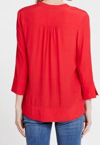 Donna che indossa una blusa rossa a maniche lunghe con pieghe sulla parte superiore della schiena e jeans blu, vista da dietro su uno sfondo bianco.