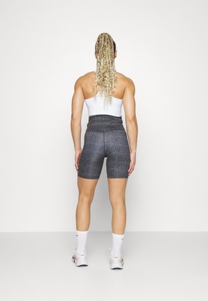 Atletisk person med blonde box fletninger iført en hvid sportstop, højtaljede grå mønstrede shorts, hvide sokker og hvide sneakers står med ryggen mod os.