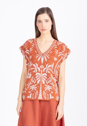 Femme portant un haut sans manches couleur orange rouille avec des motifs blancs de palmiers tropicaux et une jupe orange rouille, sur un fond blanc.
