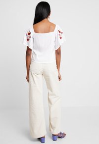Blusa blanca con mangas cortas y abullonadas que presenta bordados de flores rojas; combinada con pantalones de pana creme de pierna ancha y sandalias moradas.