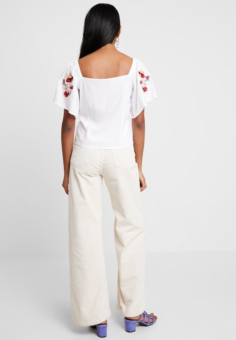 Blusa blanca con mangas cortas y abullonadas que presenta bordados de flores rojas; combinada con pantalones de pana creme de pierna ancha y sandalias moradas.