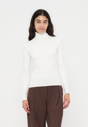 Femme aux longs cheveux foncés portant un pull à col roulé blanc et un pantalon marron à fines rayures, debout devant un fond clair uni.