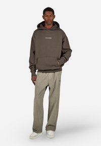 Άντρας με σκούρο καφέ φούτερ με κουκούλα που έχει την επιγραφή "PEGADOR", μπεζ καμπαρντίνες και λευκά αθλητικά παπούτσια, στέκεται με το ένα χέρι στην τσέπη.