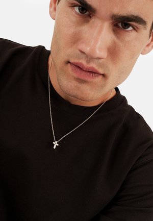 Collana a croce d'argento su una sottile catena, indossata sopra una maglietta nera a girocollo. Primo piano delle caratteristiche facciali; espressione neutra.