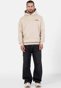 Sudadera con capucha beige con bolsillo frontal, combinada con pantalones vaqueros de pierna ancha negros con detalles desgastados y zapatillas beige.