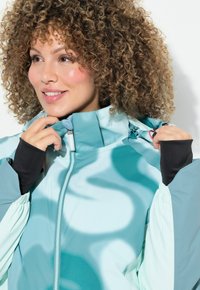Teal a světle modrá zipová bunda s vysokým límcem, elastickými manžetami a vlnkovými vzory. Materiál působí lehce a strukturovaně.