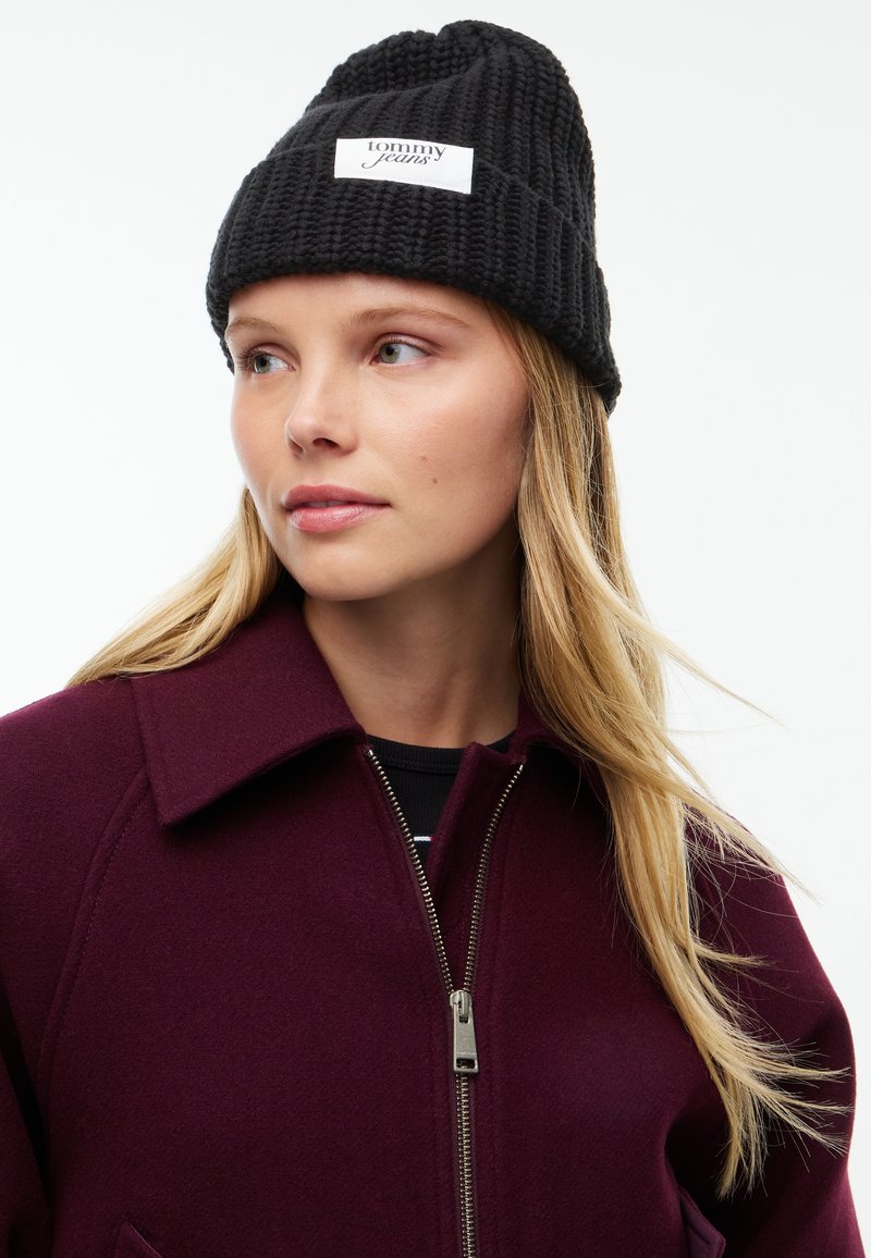 Jeune femme portant un bonnet en tricot noir Tommy Jeans et un manteau violet foncé zippé, regardant légèrement sur le côté.