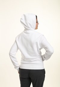 Biała hoodie, miękka w dotyku, z ribbowanymi mankietami i dołem, w luźnym kroju. Model ma na sobie ciemnoszare spodnie.