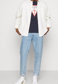 Camicia bianca a bottoni sopra una t-shirt blu navy con logo, abbinata a jeans chiari a vestibilità rilassata e sneakers bianche con dettagli scuri.
