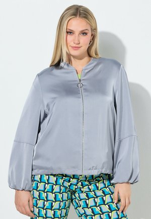 Junge Frau, die eine hellgraue Jacke mit Reißverschluss und Ballonärmeln sowie gemusterte blaue, grüne und weiße Hose trägt und vor einer weißen Wand steht.