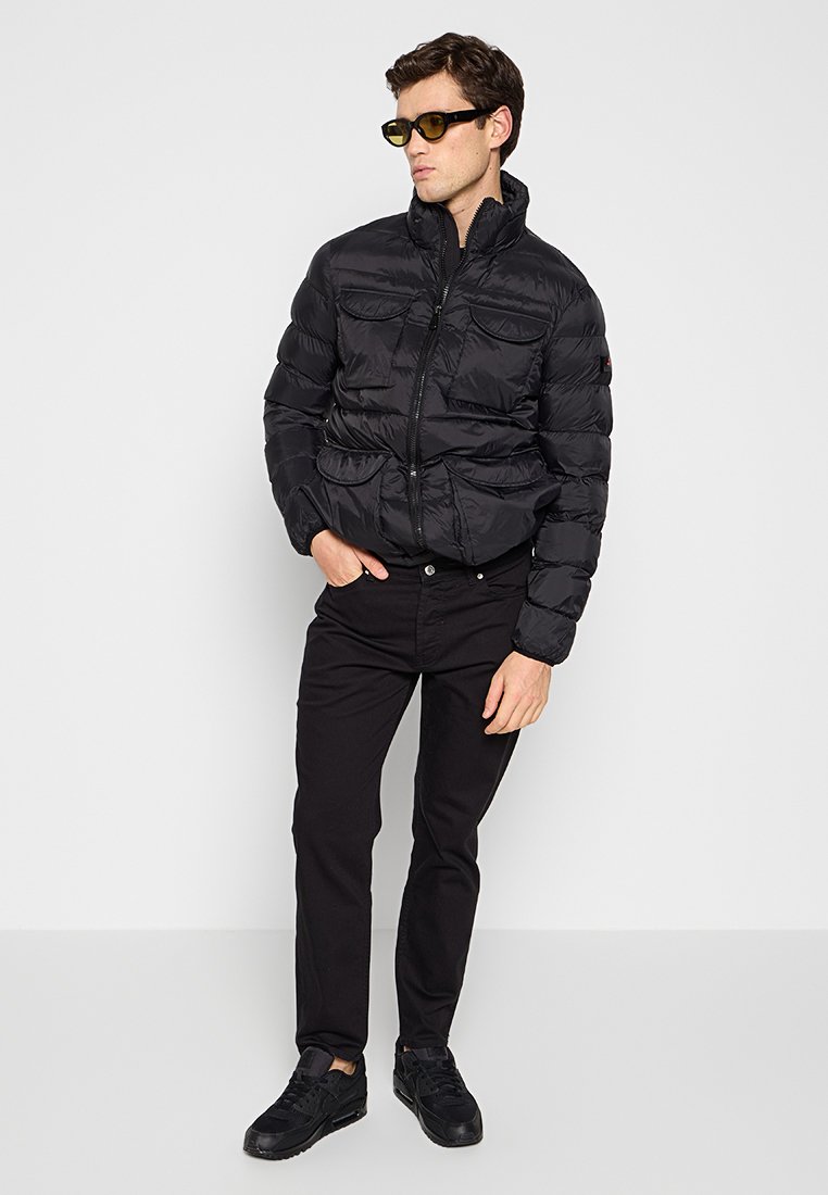 Man in een zwarte pufferjack, zwarte broek, zwarte sneakers en geelgetinte zonnebril, staand tegen een plain witte achtergrond.