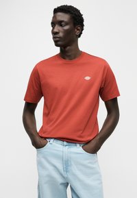 Κόκκινο βαμβακερό t-shirt με στρογγυλή λαιμόκοψη και κοντά μανίκια, με μικρό κεντημένο λογότυπο στο στήθος, συνδυασμένο με ανοιχτόχρωμο μπλε τζιν.