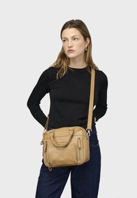 Borsa a mano in pelle color cuoio con doppi manici superiori, tracolla regolabile, tasche con cerniera, texture liscia e marchio minimale.