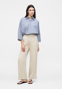 Γυναίκα που στέκεται, φοράει μπλε και λευκό ριγέ φαρδιά πουκαμίσα, μπεζ παντελόνια με φαρδιά πόδια και μαύρα σανδάλια flip-flop σε απλό φόντο.