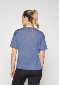 Kék, kerek nyakú, rövid ujjú póló, enyhén átlátszó anyagból készül, finom textúrával, feketére szabott leggings-szel párosítva.