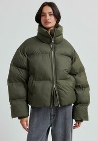 Olivgrön puffer-jacka med hög krage, kort design och silverfärgad dragkedja. Har en kviltad textur och vida ärmar.