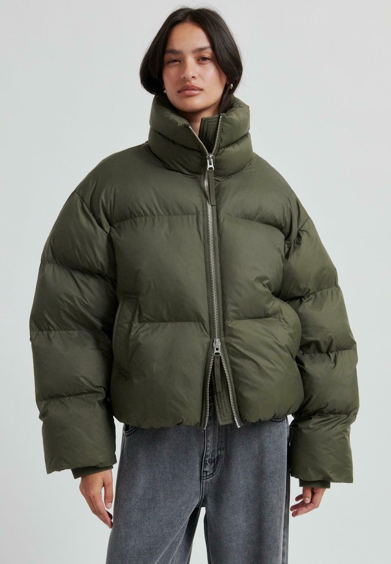 Olivgrön puffer-jacka med hög krage, kort design och silverfärgad dragkedja. Har en kviltad textur och vida ärmar.