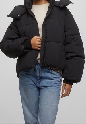 Personne portant une doudoune noire sur une chemise beige et un jean bleu clair, ajustant la fermeture éclair de la doudoune avec une main.