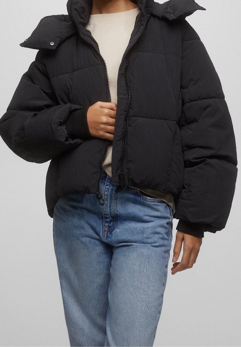 Personne portant une doudoune noire sur une chemise beige et un jean bleu clair, ajustant la fermeture éclair de la doudoune avec une main.