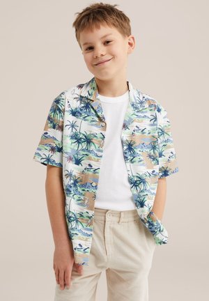 Chlapec oblečený v krátkosrstovej košeli s tropickým vzorom cez biele tričko a béžové nohavice, stojí s jednou rukou v vrecku, usmieva sa.