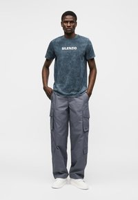 Homme portant un t-shirt bleu délavé avec l'inscription "SILENZIO", un pantalon cargo gris et des baskets blanches, debout les mains dans les poches sur un fond uni.