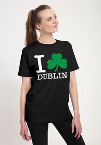 Žena s hnědým ohonem, oblečená v čern�ém tričku s bílým písmem "I", zeleným trojlístkem a bílým textem "DUBLIN" na přední straně.