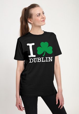 Vrouw met een bruine paardenstaart die een zwart T-shirt draagt met een witte "I", een groene klaver en de tekst "DUBLIN" in het wit op de voorkant.