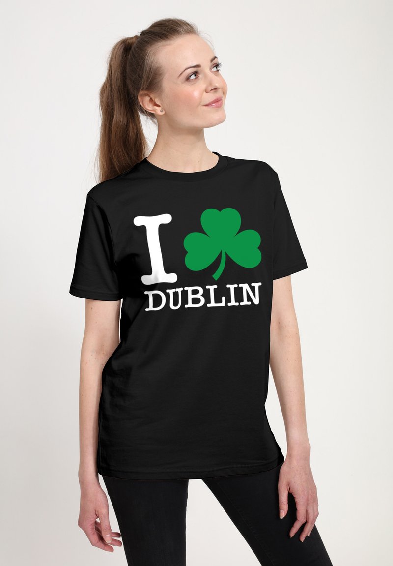 Žena s hnědým ohonem, oblečená v černém tričku s bílým písmem "I", zeleným trojlístkem a bílým textem "DUBLIN" na přední straně.