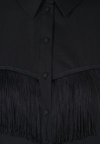Blouse noire avec un col classique, fermeture à boutons sur le devant et des détails en frange noire décoratifs au niveau de la poitrine.