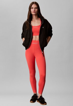 Femme debout portant des leggings corail Calvin Klein et un soutien-gorge de sport, une veste zippée noire et des baskets noires sur un fond gris uni.