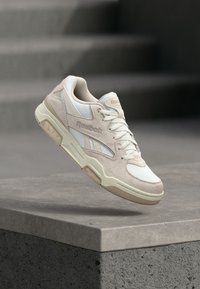 Béžové a biele tenisky Reebok s šnúrkami, detailmi strednej podrážky a logom, umiestnené nad betónovým okrajom so schodiskom v pozadí.