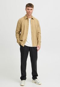 Camisa de color marrón claro con un patrón de rayas verticales, con botones al frente, llevada encima de una camiseta blanca, combinada con pantalones negros y zapatillas blancas.