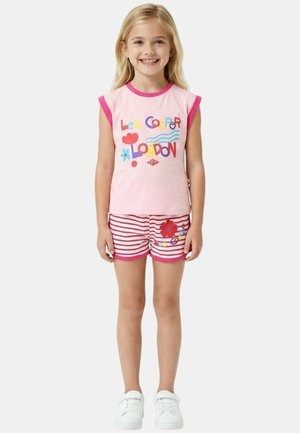 Fille souriante portant un haut sans manches rose avec le texte coloré "Lee Cooper London" et un short rose à rayures avec un motif floral ainsi que des baskets blanches.