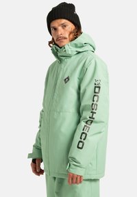 Mężczyzna w miętowej kurtce i spodniach DC Shoes z założonym kapturem i czarną czapką beanie, lekko zwrócony bokiem w stosunku do jednolitego tła.