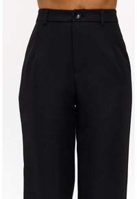 Pantalons noirs ajustés fabriqués en tissu lisse, avec un devant plat, des poches latérales et une fermeture à un bouton à la taille.