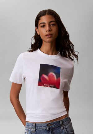 Giovane donna con capelli mossi che indossa una t-shirt bianca Calvin Klein con una grafica sfocata di un fiore rosso e jeans casual blu.