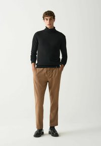 Črna turtleneck pulover, bež hlače in črne čevlje. Ožji kroj, gladka tekstura, z žepi in subtilnimi oblikovalskimi podrobnostmi.