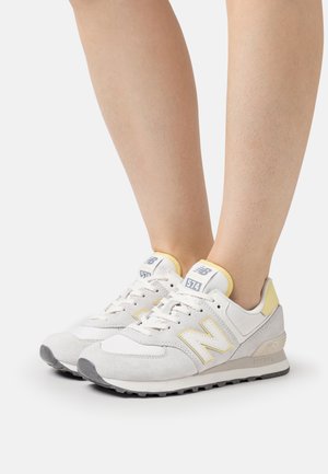 Weiße und hellgelbe New Balance 574 Sneakers an hellhäutigen Füßen, vor einem schlichten weißen Hintergrund gezeigt.