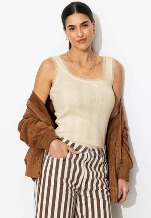 Femme portant un haut beige sans manches avec bordure en dentelle, un pantalon rayé marron et une veste marron posée sur ses épaules, souriant à la caméra.