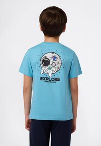 Niño vistiendo una camiseta de color azul claro con un astronauta de dibujos animados sosteniendo una bandera en la luna y el texto "EXPLORA MÁS ALLÁ DE LA LUNA" en la parte de atrás.