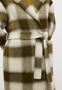 Cappotto di lana a scacchi marrone e crema con colletto a scialle, tasche frontali e una cintura abbinata annodata in vita. Texture soffice.